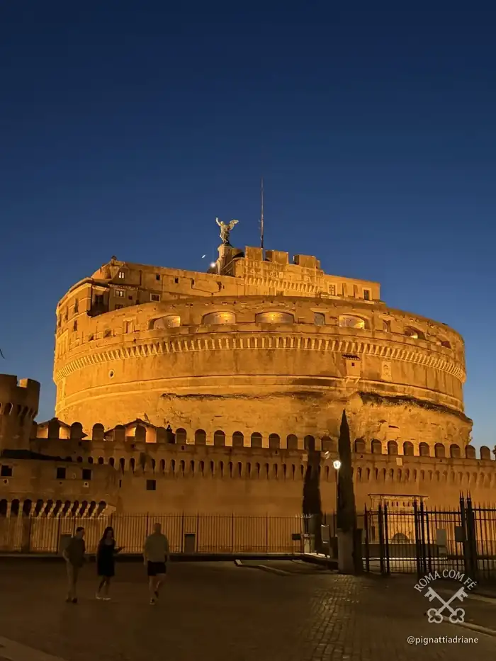 O castelo visto do caminho ribeirinho, abaixo do Lungotevere, com os refletores já acesos. De dia ele é um cilindro de tijolo; à noite fica dourado.