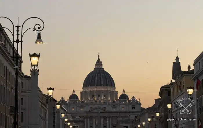 A Via della Conciliazione ao anoitecer. É por aqui que a cúpula aparece de longe, no lugar onde o terceiro braço da colunata deveria fechar a praça.